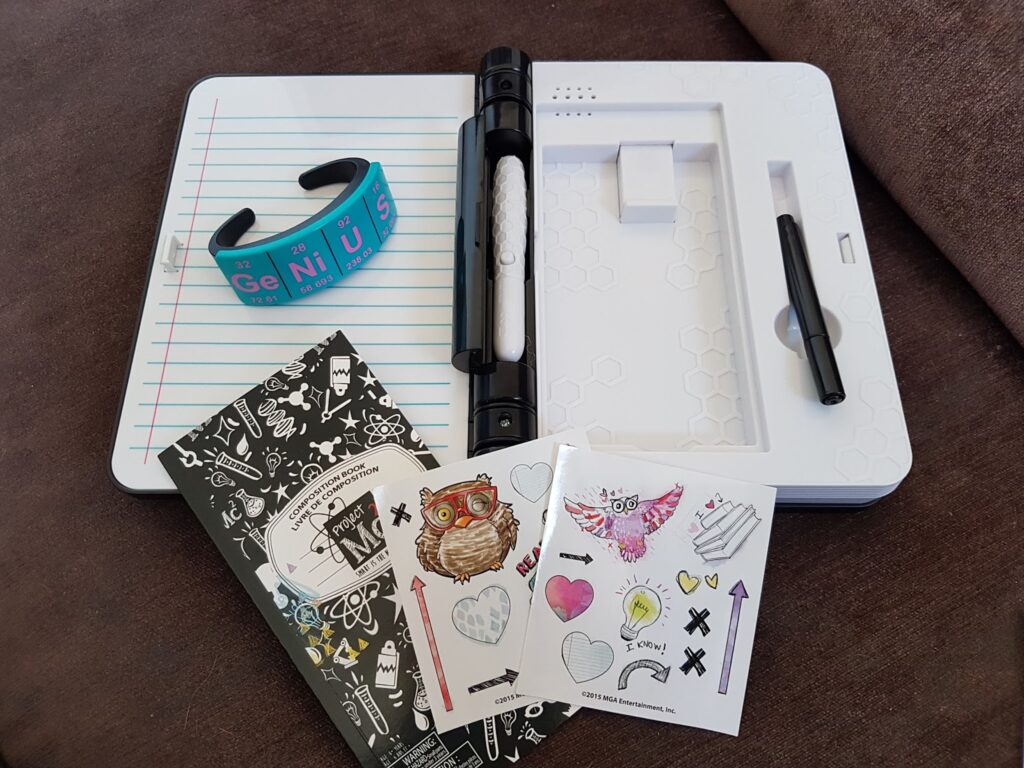 An open notebook displays lined paper on the left and a white plastic section holding a black pen on the right. In front are two sticker sheets, a black-and-white patterned booklet, and a blue wristband labelled "GENIUS." Items are on a brown surface.