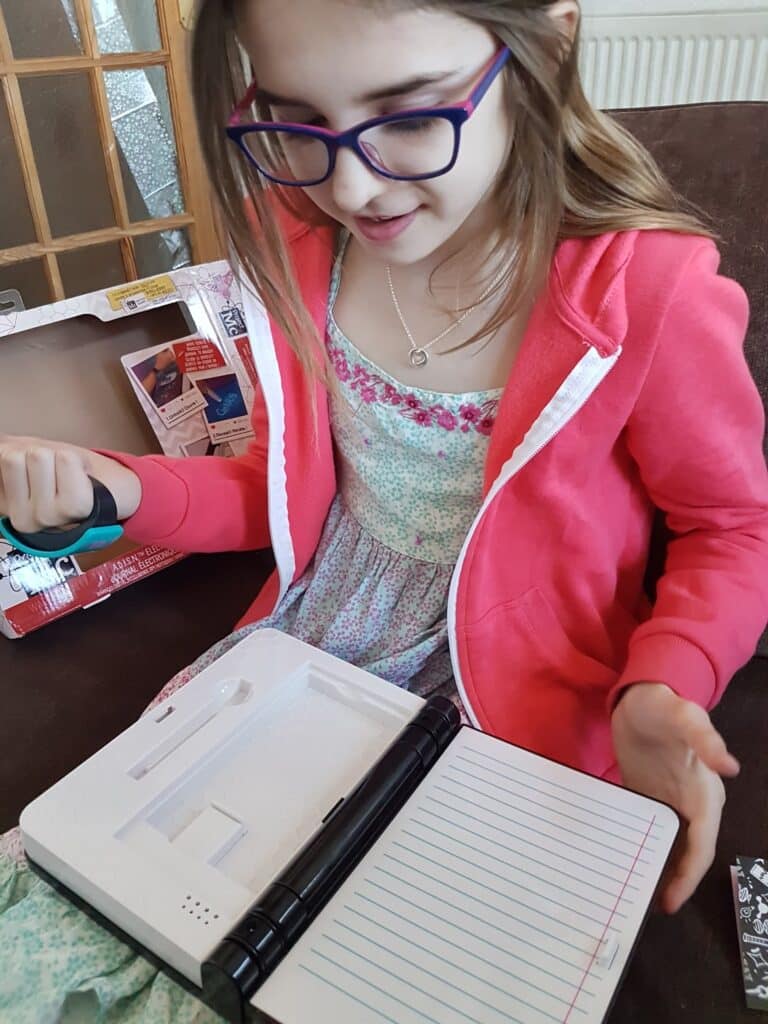 A young girl with glasses and a pink hoodie sits on a sofa, looking at an open electronic notebook with lined pages. She holds a pen above the notebook and appears to be engaged and curious. An opened box is visible beside her.