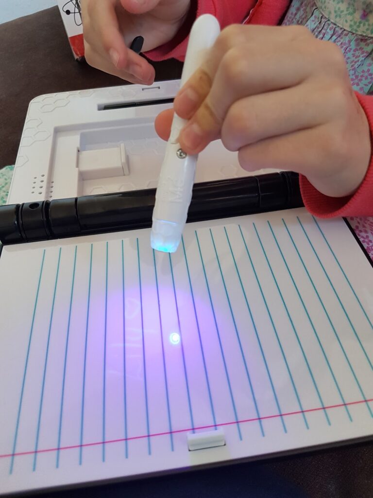 A child holds a white pen with a blue LED light, writing on a blank, lined white notebook. The blue light reveals invisible ink on the page. The child wears a red jumper and a patterned shirt. The background shows part of a plastic case.