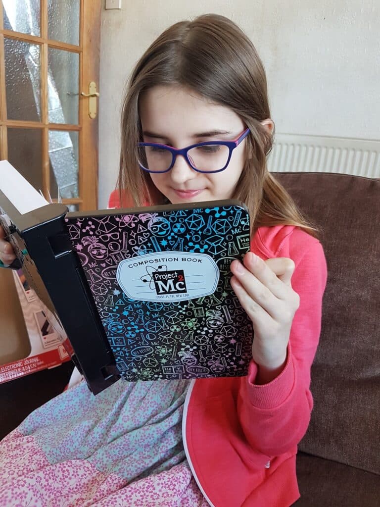 A young girl with long brown hair and purple glasses sits on a sofa, looking down and reading a black "Project MC2" exercise book with colourful science-themed doodles on the cover. She wears a pink jumper over a patterned dress.