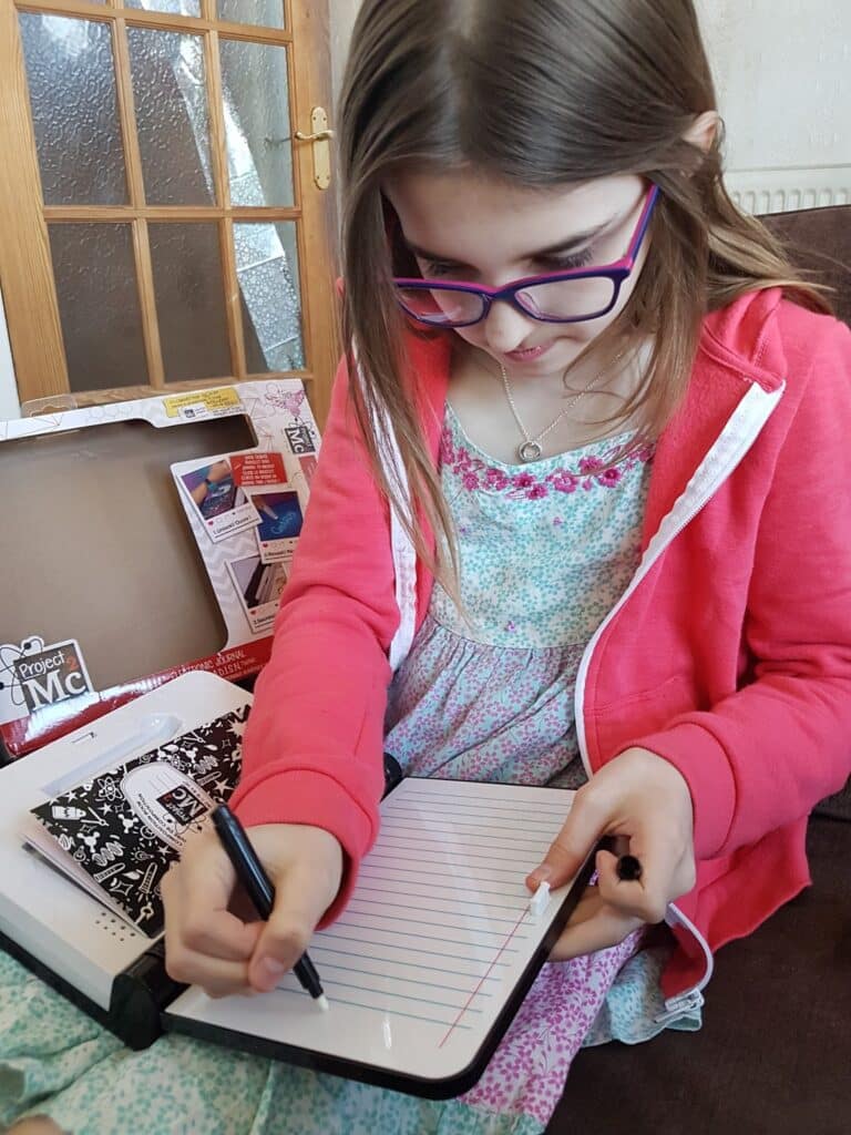 A young girl with long brown hair and purple glasses is sitting on a sofa, wearing a pink hoodie over a floral dress. She is writing on a small whiteboard with a black marker. A Project MC2 science kit box is open beside her.