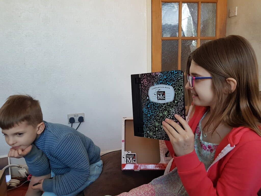 A young girl with glasses and long brown hair, wearing a pink hoodie, holds up a patterned notebook and smiles. Next to her, a young boy in a blue jumper leans forward with his chin resting on his hand. They are indoors near a wooden door.