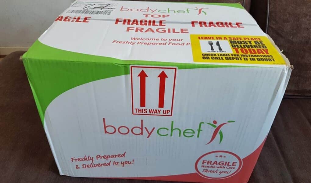A large cardboard box labelled "bodychef" sits on a brown sofa. The box is white, green, and red, marked "FRAGILE" and "THIS WAY UP" with red arrows. Instructions and delivery notes are printed on the top and side, emphasising care and freshness.
