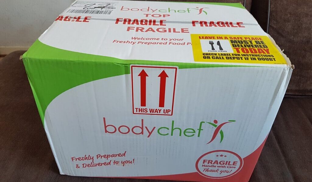 A large cardboard box labelled "bodychef" sits on a brown sofa. The box is white, green, and red, marked "FRAGILE" and "THIS WAY UP" with red arrows. Instructions and delivery notes are printed on the top and side, emphasising care and freshness.