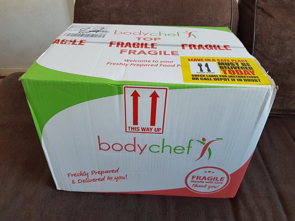 A large cardboard box labelled "bodychef" sits on a brown sofa. The box features red and green branding, "FRAGILE" warnings, "THIS WAY UP" arrows, and delivery instructions. The text indicates it contains freshly prepared food.
