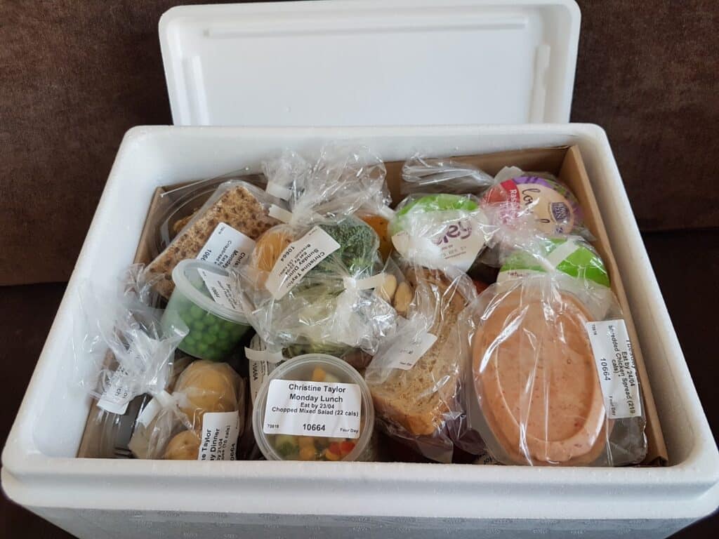 A white polystyrene box filled with neatly packed and labelled food portions, including bread, cold meat, peas, cheese, fruit, and various meal containers. Each item is in clear plastic or lidded containers, organised for easy identification and distribution.