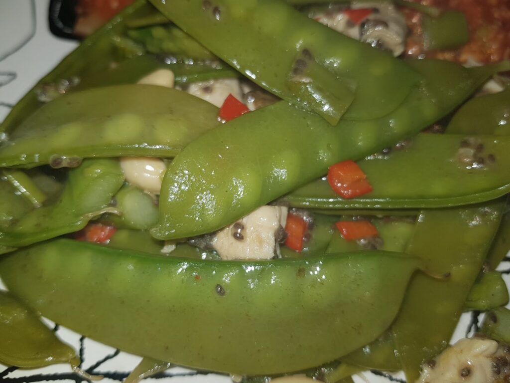 Close-up of cooked mangetout mixed with chopped red pepper, mushrooms, and black pepper seasoning. The vegetables appear glossy and are served on a white plate with a hint of another dish visible in the background.