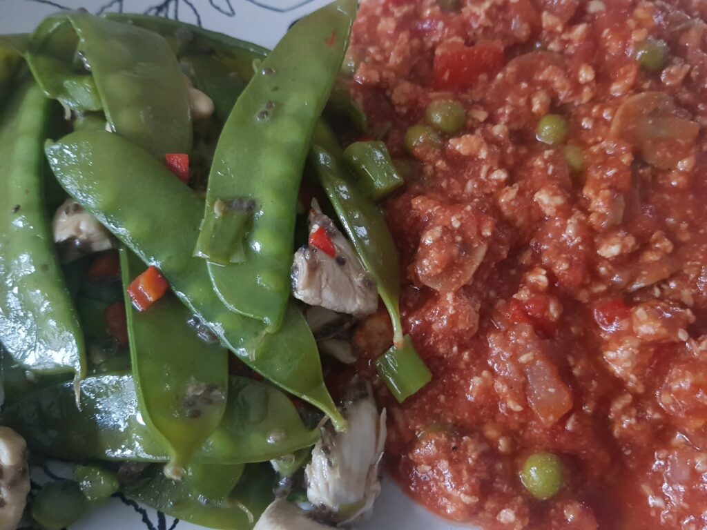 A plate with two sections: on the left, stir-fried mangetout with mushrooms and red peppers; on the right, a chunky tomato-based sauce with peas and minced meat. The vegetables appear glossy, and the sauce has a reddish hue.