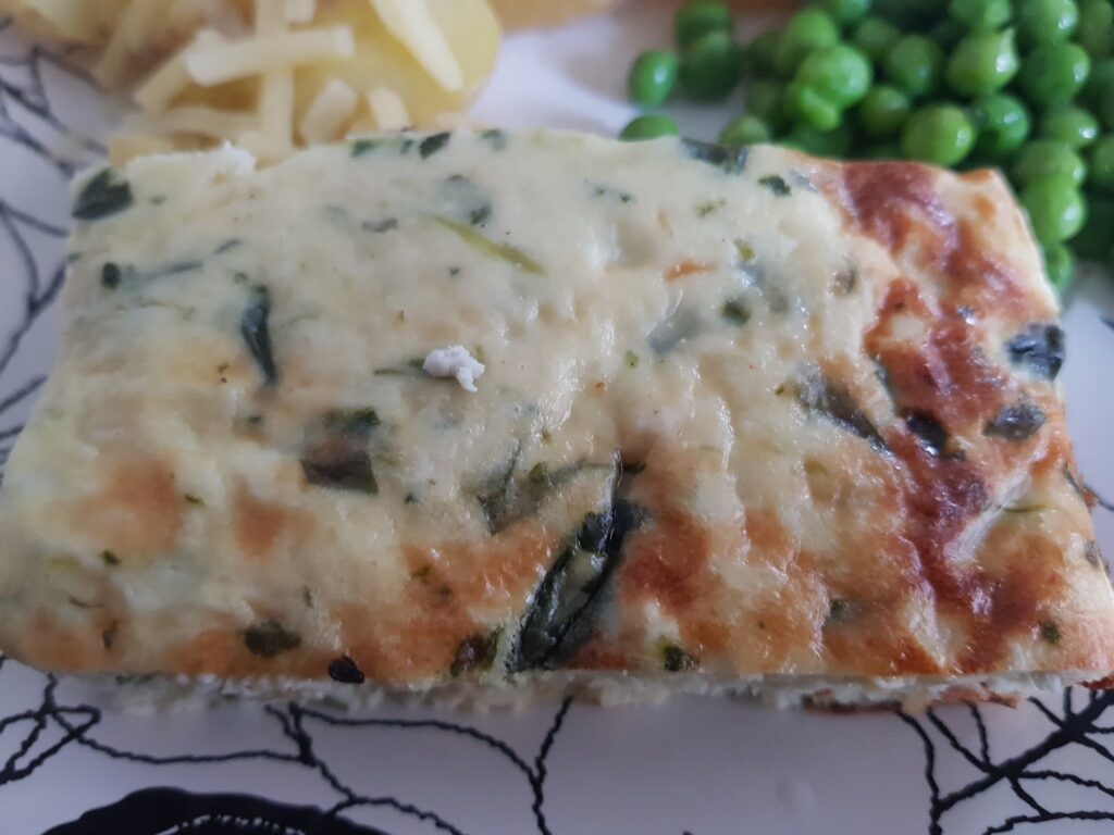 A close-up of a rectangular slice of baked spinach and cheese casserole with a golden-brown top. The casserole is soft with visible pieces of spinach. In the background are green peas and a small portion of grated cheese on a patterned plate.