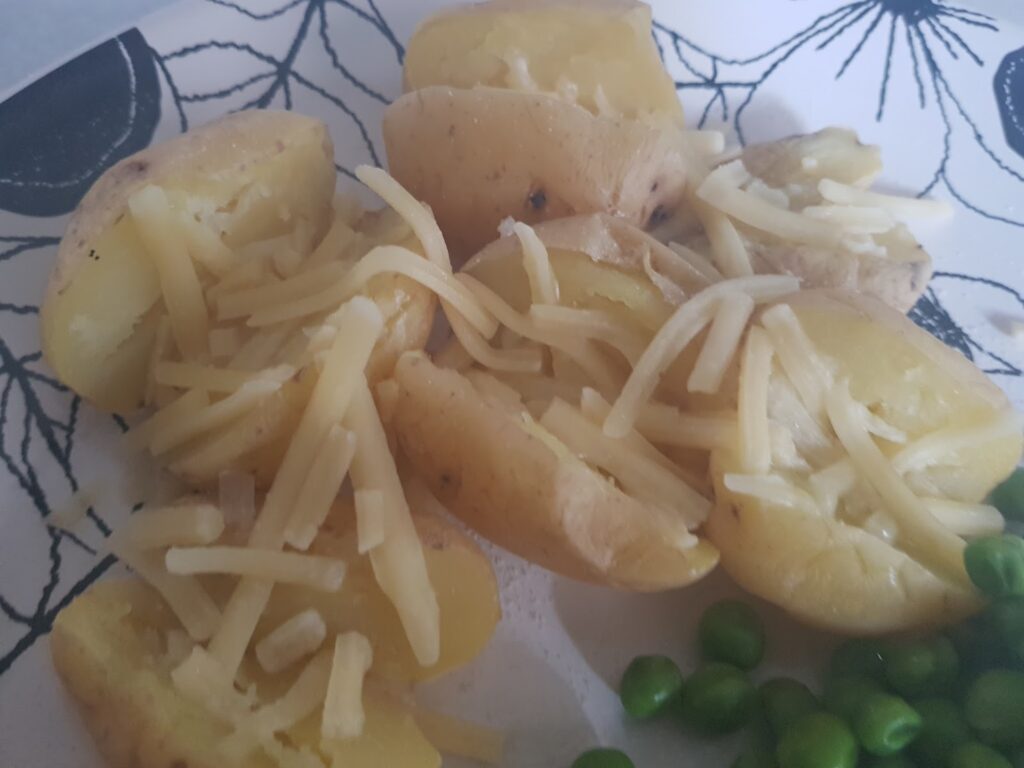 A close-up of boiled potatoes cut in half and sprinkled with grated cheese, served on a white plate with a black leaf design. Green peas are visible at the edge of the plate. The meal appears simple and home-made.