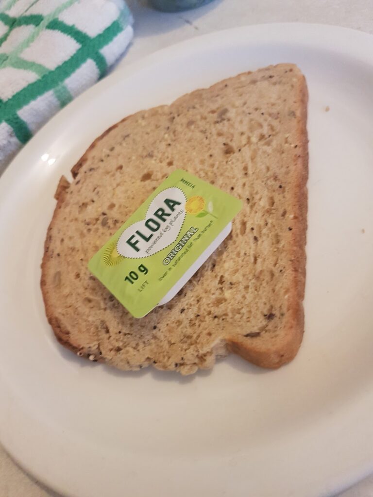 A single slice of brown bread sits on a white plate. On top of the bread is a small, unopened tub of Flora margarine (10g). A green and white chequered cloth is partially visible in the background.