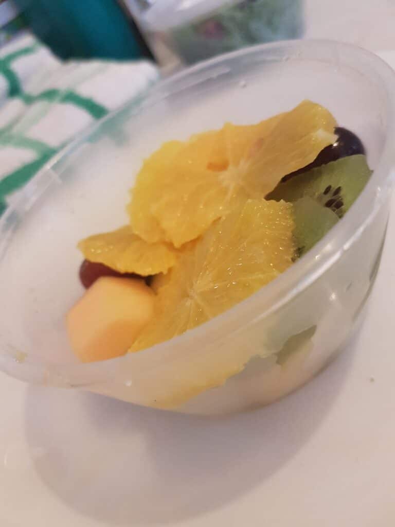 A clear plastic bowl filled with assorted fruit pieces, including orange segments, kiwi slices, red grapes, and a yellow melon chunk, sits on a white surface with a green-and-white cloth slightly visible in the background.