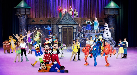 A large group of Disney characters, including Mickey, Minnie, Donald, Goofy, princesses, Olaf, and characters from Finding Nemo, perform together in colourful costumes on an ice rink in front of a fairytale castle set with purple lighting.