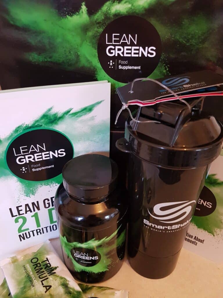 A display featuring Lean Greens supplements, including a large bottle, sachets, and a brochure titled "Lean Greens 21 Day Nutrition." A black SmartShake shaker bottle is in the foreground, with green branding elements and a leafy background.