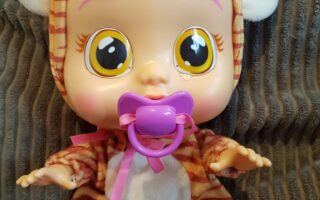 A large-eyed baby doll with pink hair and a purple dummy sits on a grey textured cushion. The doll wears a tiger-striped babygrow with white ears and paws, and has a pink bow tied to the dummy. Its arms and legs are slightly spread.