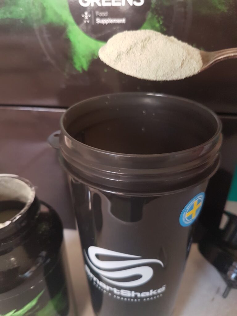 A spoonful of green powder is held above a black shaker bottle labelled “SmartShake.” An open container of powder sits nearby, and a box labelled “Greens Food Supplement” is visible in the background.