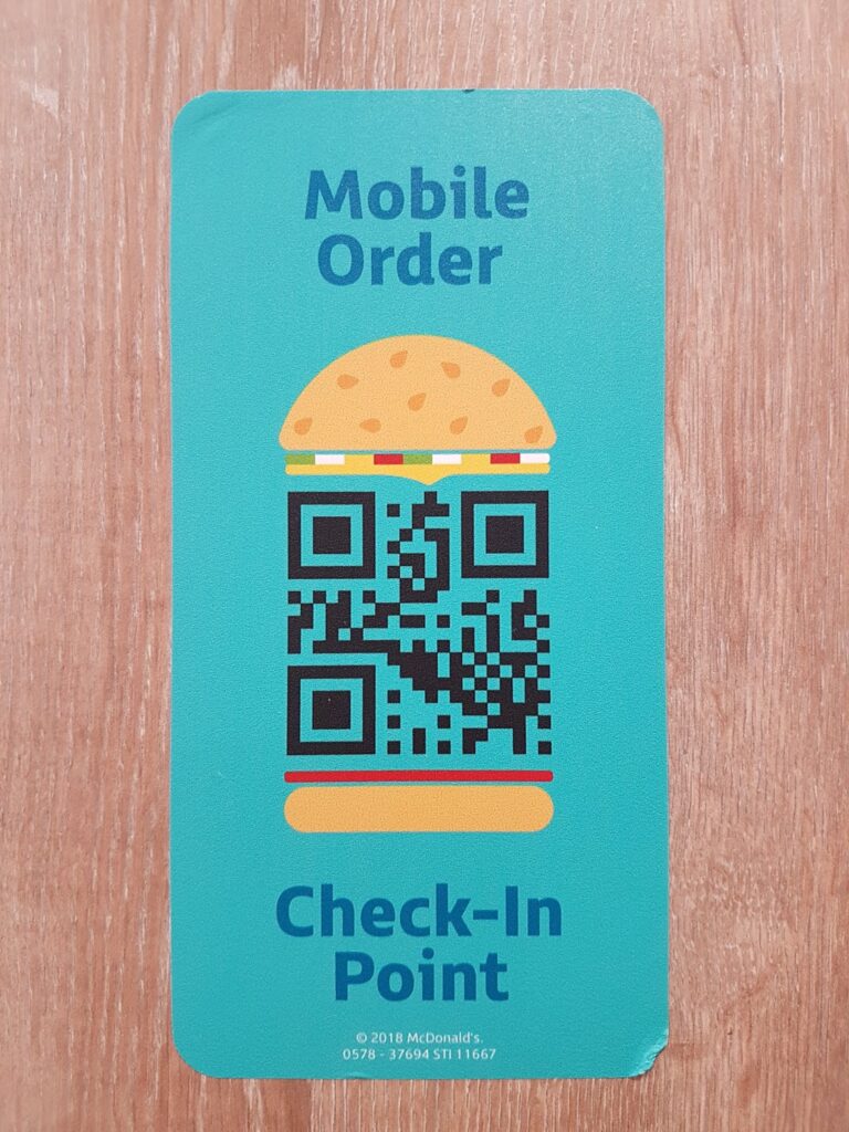 A teal card on a wooden surface displays a burger-shaped QR code. Above, blue text reads "Mobile Order." Below, blue text reads "Check-In Point." Small copyright and numbers appear at the bottom.
