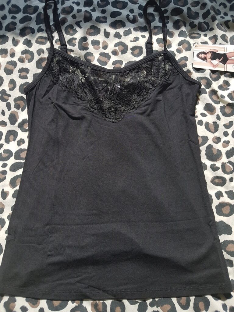 A black camisole with thin straps and lace trim at the neckline is laid flat on a leopard-print fabric. A product tag with an illustrated woman in lingerie is seen in the top right corner. The camisole appears new and unworn.