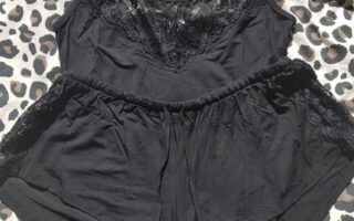 A black camisole and matching shorts pyjama set with lace trim is laid flat on a leopard-print fabric. The set is sleeveless, and a tag with a photo of a model wearing the outfit is attached in the upper right corner. Sunlight creates soft shadows on the set.