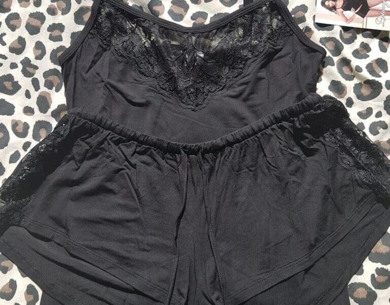 A black camisole and matching shorts pyjama set with lace trim is laid flat on a leopard-print fabric. The set is sleeveless, and a tag with a photo of a model wearing the outfit is attached in the upper right corner. Sunlight creates soft shadows on the set.