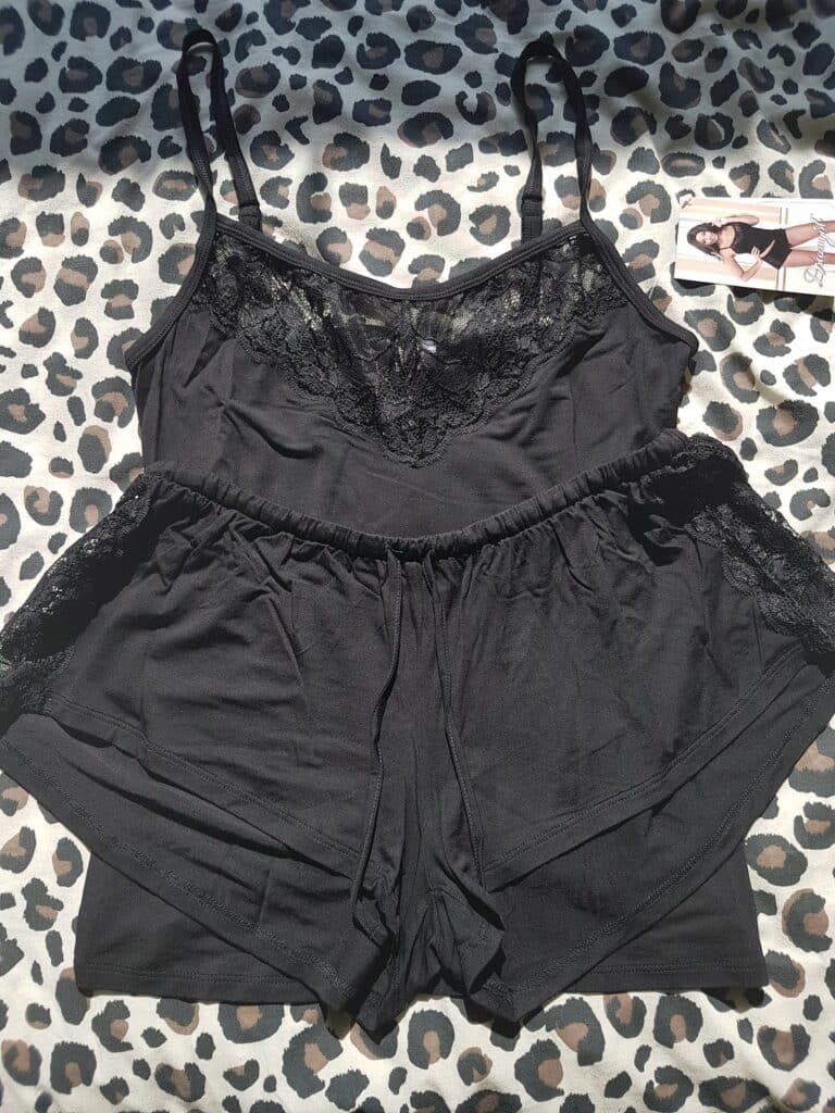 A black camisole and shorts pyjama set with lace trim on the top and sides, displayed on a leopard print fabric. A tag with a model wearing the set is visible to the upper right of the clothing. Sunlight shines across the outfit.