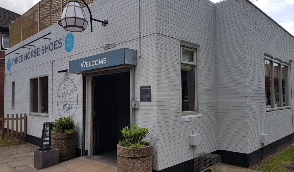 A white brick building with a sign reading “Three Horse Shoes” and “Welcome” above the entrance. Outdoor planters flank the door, and a chalkboard sign sits on the pavement. Large windows line the side, and a hanging lantern decorates the front.