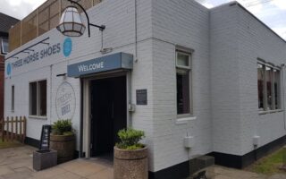 A white brick building with a sign reading “Three Horse Shoes” and “Welcome” above the entrance. Outdoor planters flank the door, and a chalkboard sign sits on the pavement. Large windows line the side, and a hanging lantern decorates the front.
