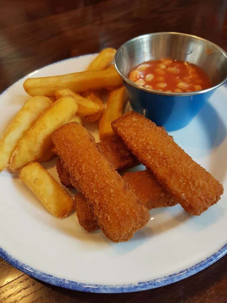 A white plate with thick-cut chips, three breaded fish fingers, and a small metal cup filled with baked beans in tomato sauce, on a dark wooden table.
