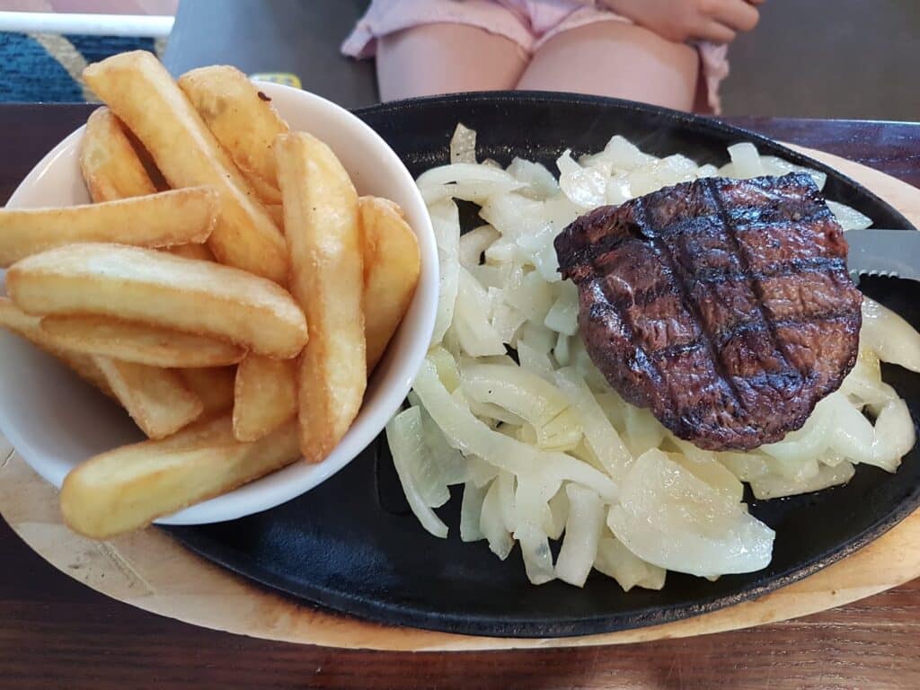 A plate with thick-cut chips in a white bowl, next to grilled steak on a bed of sautéed onions, served on a black platter. In the background, a person’s crossed legs in pink shorts are partially visible.
