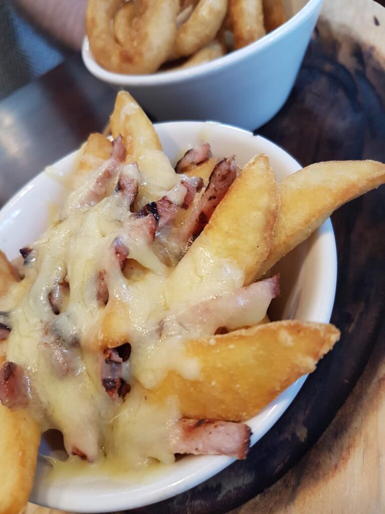 A white bowl contains thick-cut potato wedges topped with melted cheese and bacon pieces. In the background, a partially visible bowl holds onion rings. Both bowls are placed on a dark wooden board.