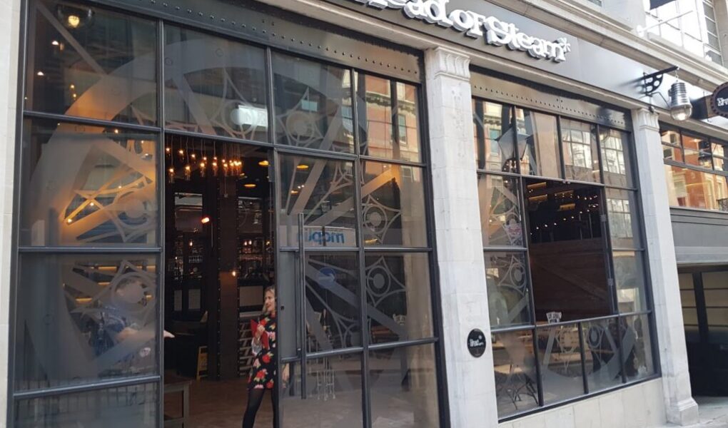 A woman in a red and black coat exits “The Head of Steam” pub, which has large glass windows, an industrial interior, and a visible clockwork gear motif inside. The exterior is modern with stone columns and a black sign with white lettering.