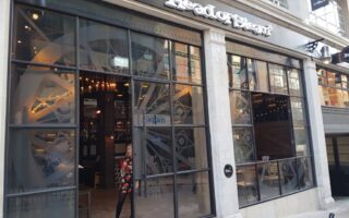 A woman in a red and black coat exits “The Head of Steam” pub, which has large glass windows, an industrial interior, and a visible clockwork gear motif inside. The exterior is modern with stone columns and a black sign with white lettering.
