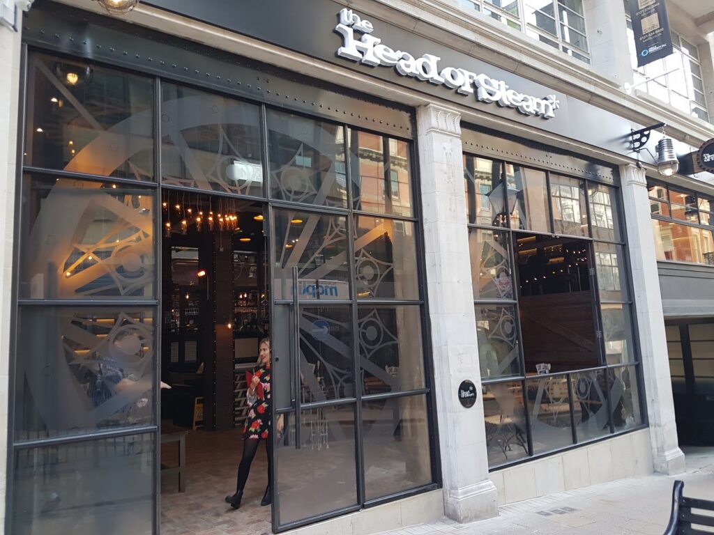 Street view of The Head of Steam bar with large glass windows, displaying a visible interior featuring industrial decor and a large gear design. A woman in a floral coat stands near the entrance, and a “We’re open” sign hangs on the door.