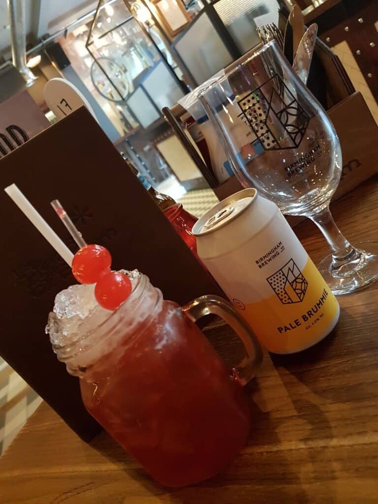 On a wooden table, there is a mason jar glass with a red iced drink topped with two maraschino cherries, a can of Pale Bruhh beer, an empty stemmed glass, menus, condiments, and cutlery in a casual restaurant setting.