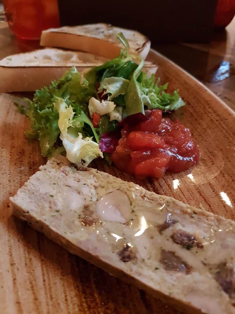 A wooden plate with a slice of terrine studded with nuts and herbs, a small fresh green salad, a serving of chunky red chutney or relish, and two slices of toasted bread in the background. The food is arranged neatly and looks appetising.