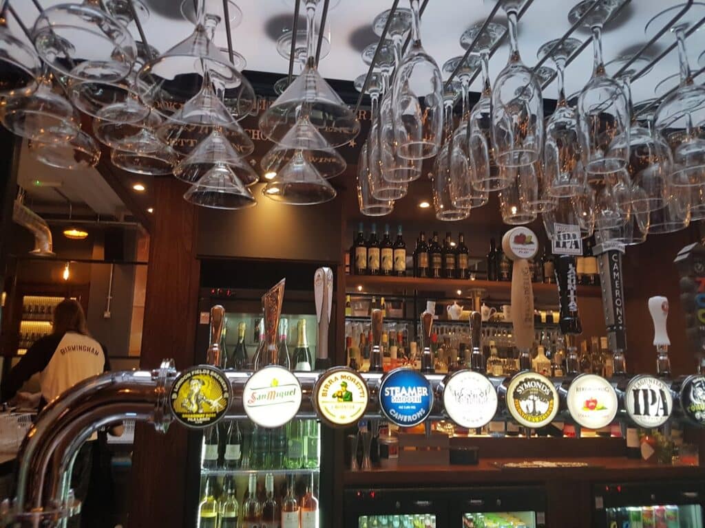 A bar with rows of glasses hanging upside down above a counter. Multiple beer taps with colourful labels are lined up, and bottles of spirits are arranged on shelves in the background. A person wearing a white shirt stands behind the bar on the left.