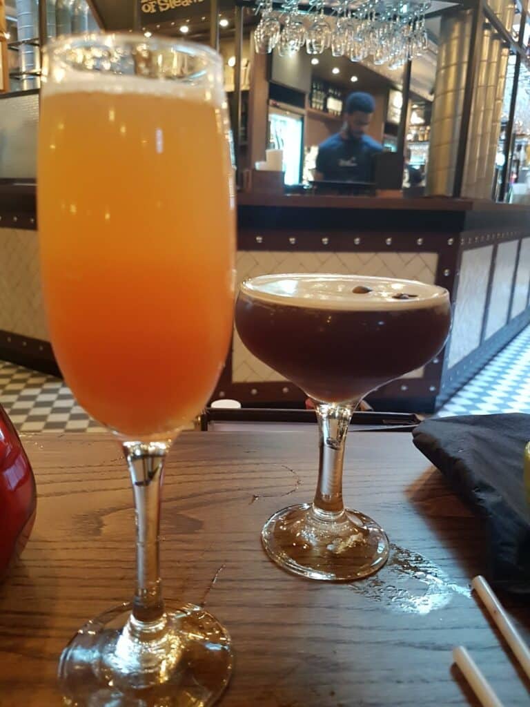 A close-up of two cocktails on a wooden table: one is orange and fizzy in a flute glass, the other is dark with frothy foam in a coupe glass. The background shows a bar area with a barman and warm lighting.