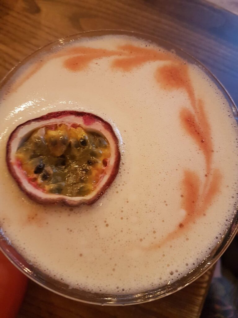 A close-up of a creamy cocktail in a glass, garnished with a halved passion fruit. The drink has a smooth, frothy surface with swirling orange streaks, and the glass is set on a wooden table.