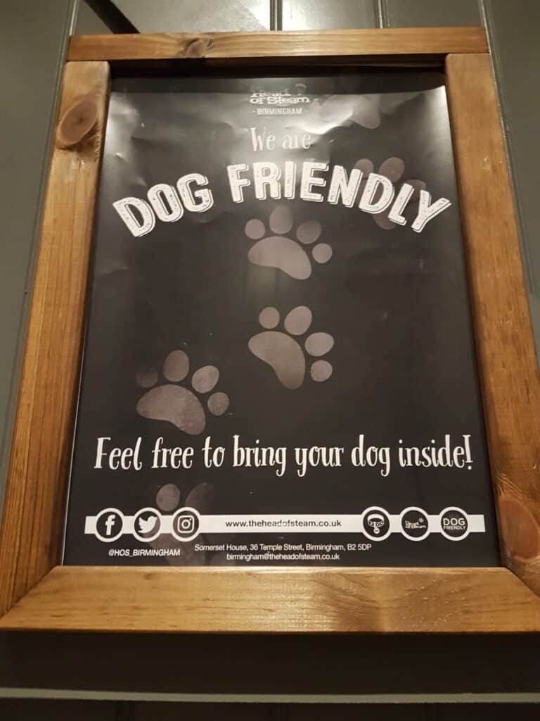 A framed sign reads: “We are DOG FRIENDLY. Feel free to bring your dog inside!” with paw prints in the background. Social media icons and contact details for The Head of Steam Birmingham appear at the bottom.