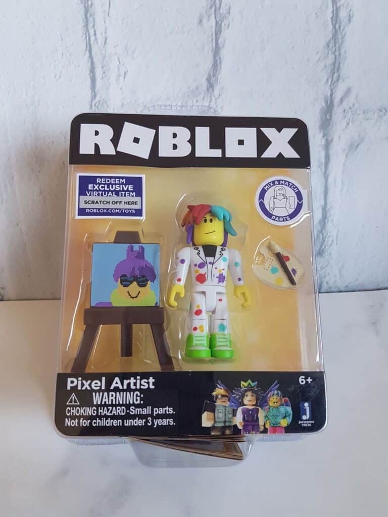 A packaged Roblox Pixel Artist toy features a figure in colourful, paint-spattered clothes, a palette, and a painted easel. The packaging has "ROBLOX" at the top, age warning, and an exclusive virtual item code sticker in the corner.