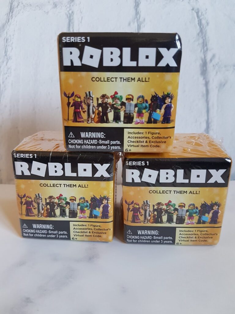 Three Series 1 Roblox mystery boxes are stacked on a light surface. Each box features images of collectable Roblox figures, a warning about small parts, and text stating each contains one figure, accessories, collector’s checklist, and a virtual item code.