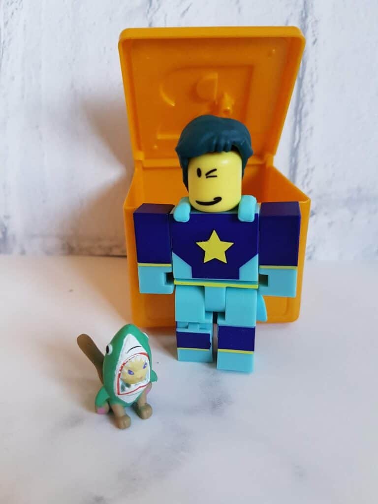 A blue and purple blocky toy figure with a yellow star on its chest stands beside a small animal figure in a green costume. Behind them is an open yellow box, all set on a white surface against a light background.