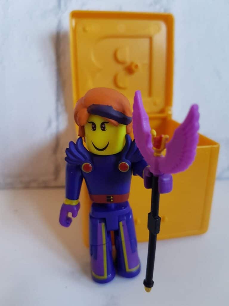 A toy figure with a yellow face, purple and yellow outfit, and orange hat stands in front of an open yellow box. The figure is smiling and holds a purple staff with wing-like shapes at the top. The background is plain and light-coloured.
