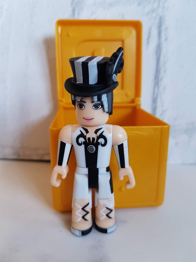 A toy figure with white skin, black-and-white outfit, and black markings stands in front of an open yellow box. The figure wears a tall black-and-white striped hat with goggles and has short black hair, black gloves, and white boots with black details.