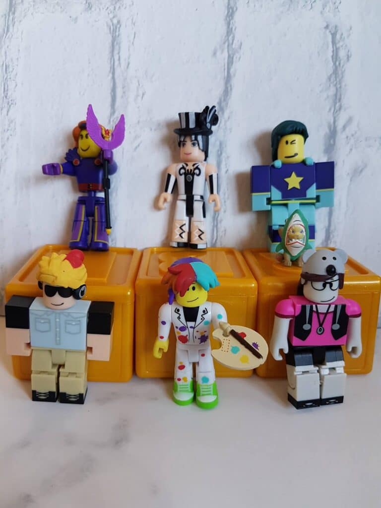 Six colourful Roblox toy figures stand on and in front of orange blocks against a white brick background. Each figure has unique outfits and accessories, such as hats, glasses, and a surfboard, showcasing different playful character designs.
