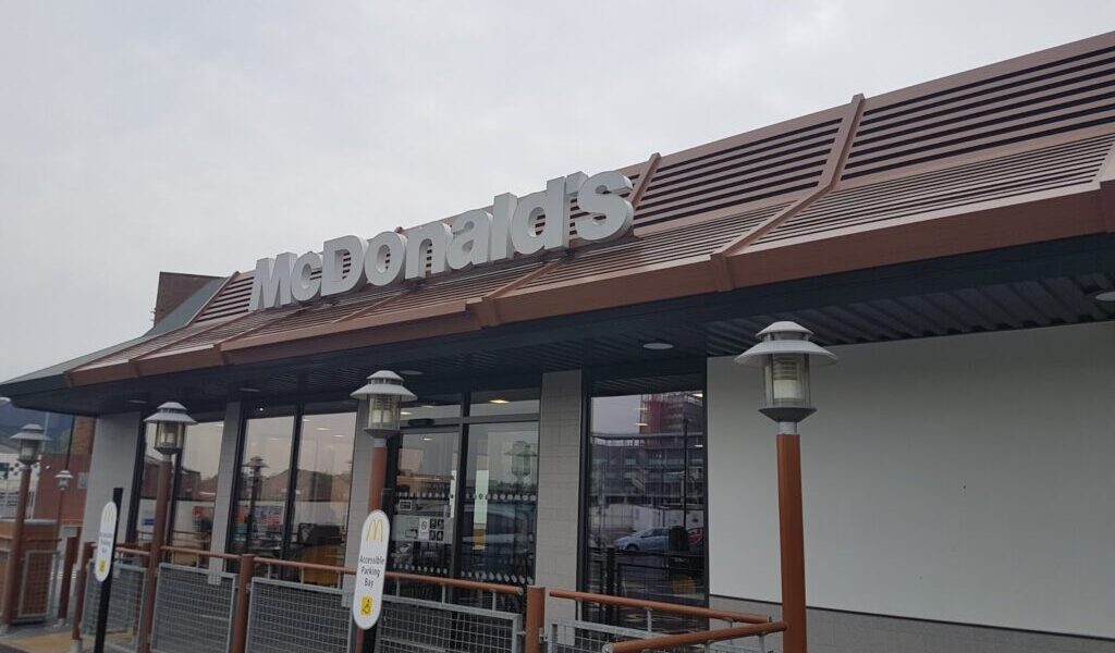 A McDonald’s restaurant exterior with a modern brown and grey design, large windows, and the McDonald’s logo sign mounted on the roof. Outdoor lights line the entrance. The sky is overcast.