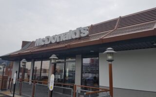 A McDonald’s restaurant exterior with a modern brown and grey design, large windows, and the McDonald’s logo sign mounted on the roof. Outdoor lights line the entrance. The sky is overcast.