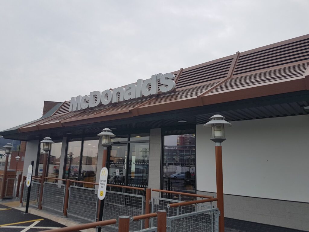 A McDonald’s restaurant exterior under a cloudy sky, featuring the large silver “McDonald’s” sign. The building has glass windows, outdoor lamps, and a modern brown and grey facade. A reflection of nearby structures is visible in the windows.