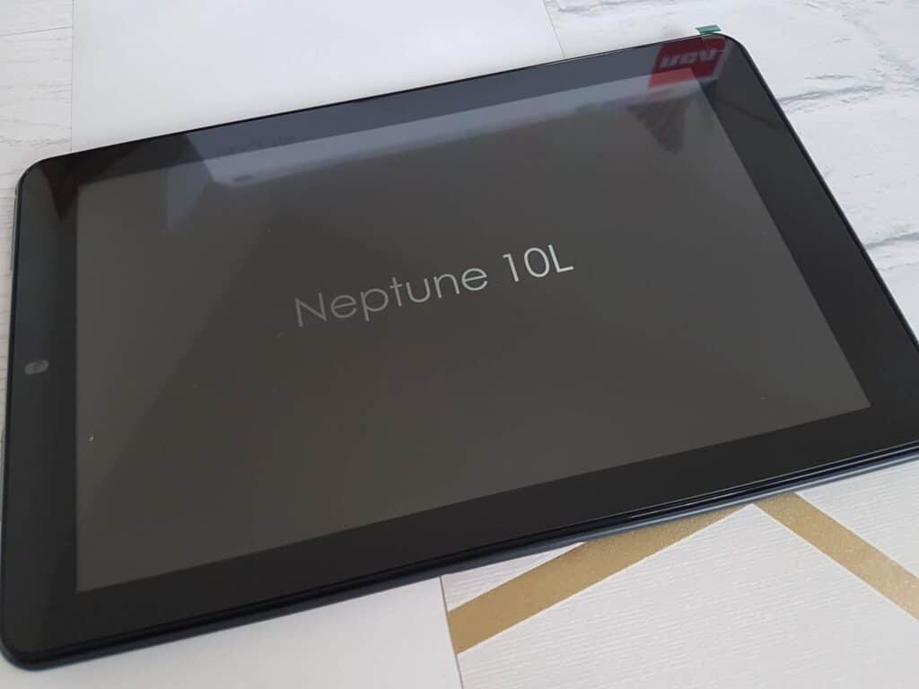 A black tablet displaying the text "Neptune 10L" on its screen lies on a white surface with gold and grey lines. Part of a red sticker is visible at the top right corner of the device.