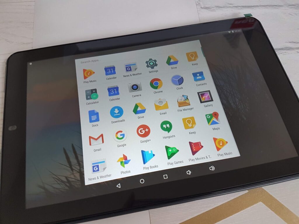 A black tablet is displayed on a white surface, showing its home screen with various app icons, including Google, Gmail, Chrome, Maps, YouTube, Play Store, Drive, Calendar, Photos, Play Music, Play Movies and others.